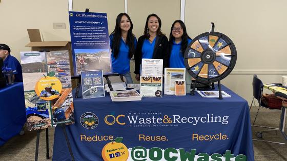 Three OC Waste & Recycling employees at vendor booth at Cool OC event. Vendor table has spinning wheel and displays.