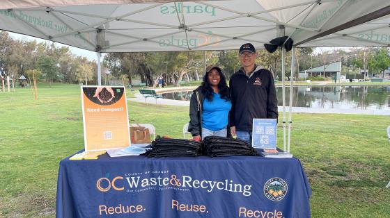 One woman and one man standing on grass behind a table with a tablecloth reading "OC Waste and Recycling""