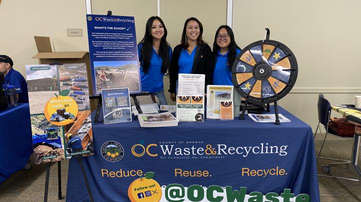 Three OC Waste & Recycling employees at vendor booth at Cool OC event. Vendor table has spinning wheel and displays.