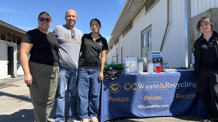 Three women and one man standing in front of a table with a tablecloth reading "OC Waste and Recycling"."