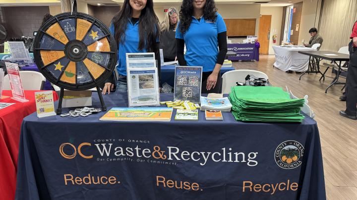 Two women standing behind a table with a tablecloth reading "OC Waste and Recycling"."